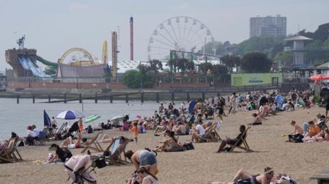 A wide picture of Southend beach with multiple people sitting on the beach, many on deck chairs. In the background is a funfair with a big wheel. It is a hazy but sunny day.