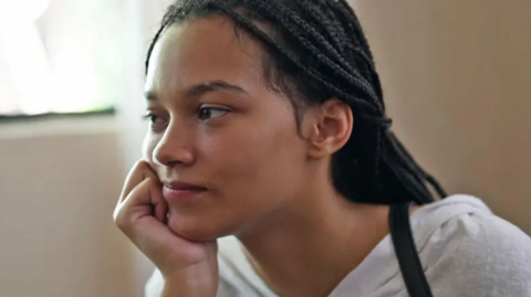 A woman sits indoors near a softly lit window, resting her face gently on one hand. Her hair is styled in neat braids, and they’re wearing a light‑coloured top that blends nicely with the warm, natural light coming in. The overall mood feels reflective and peaceful