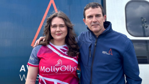 Two people standing in front of a train smiling. A woman with long brown curly hair wearing a bright pink top which says Molly Ollys on it. The man has short brown hair and is also smiling. He has his arm around his daughter. He is wearing a navy jacket with avanti west coast on it. 