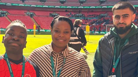 Kayden Douglas-Imasuen, his mum Lizzy and Aubaid Aftar all standing by the side of the pitch at Wembley stadium. They all have lanyards on. Kayden has on a Luton football shirt, Lizzy, her hair tied back and a black and white striped shirt, and Aubaid a black gilet and green hoodie. There are lots of red seats behind them and supporters sitting in some of them. A photographer is behind them on the pitch.