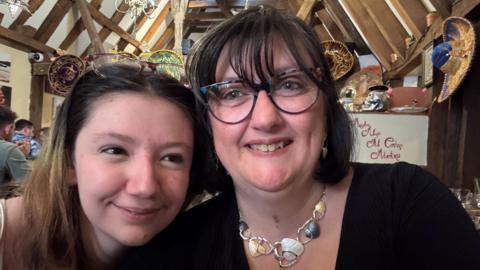 Lilimae (left) and Tracey smile as they huddle together. They are pictured inside a public building with exposed wooden beams.