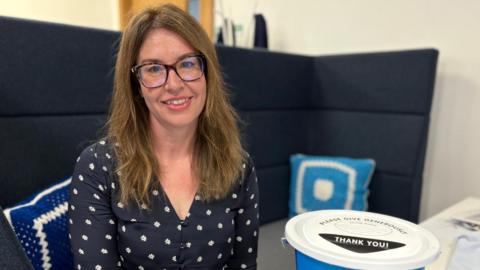 Rachel is sat on a high-backed navy chair in an office, surrounded by Diabetes UK promotional material. She is wearing a navy, floral blouse. She has long brown hair and is wearing dark-rimmed glasses and is smiling at the camera.