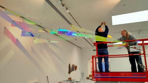 Two technicians, a man and a woman, on a red hydraulic platform in a white museum room. They are putting the final pieces to a glass artwork which hangs from the ceiling. It is a series of glass triangles in different colours which reflect on to the white walls. 