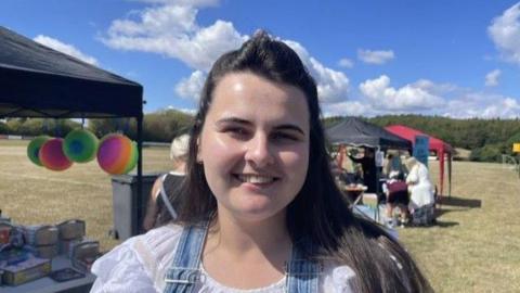 Chloe Fogg pictured at the event with long brown hair and wearing blue denim dungarees and a white top. Behind her are stalls and people gathered around them.