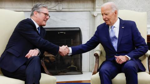 Keir Starmer, on the left wearing a navy suit, sits on a cream armchair shaking hands with Joe Biden, on the right, also in a cream armchair, wearing a lighter shade of navy. There is a marble fireplace between them and ornate wallpaper on the walls