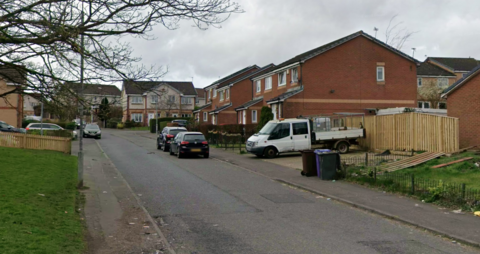 Residential street with brick semi-detached houses, parked cars along the road, a white van in a driveway, wooden fencing, and an overcast sky