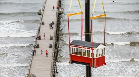 A red and white wooden carriage with a sloped roof being lifted by crane over a pier and sea. People are on the pier and the sea has waves.