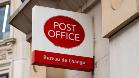 A still image of a red and white Post Office sign attached to a white stone building. Underneath the logo, which is a red oval with Post Office written in white letters, reads 'Bureau de Change'