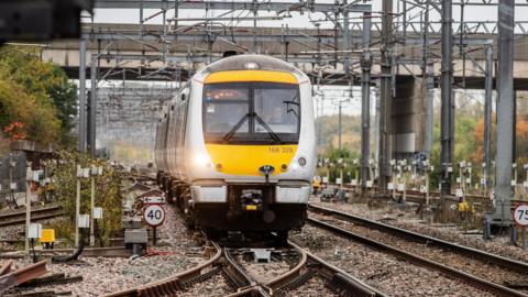 A yellow and silver train running on a railway track on a grey overcast autumnal day. Two members of staff sit in the front of the cabin, one is driving the train.