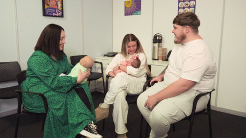Two parents with their baby sit in a semi-circle with a family nurse, who is demonstrating something using a model baby.