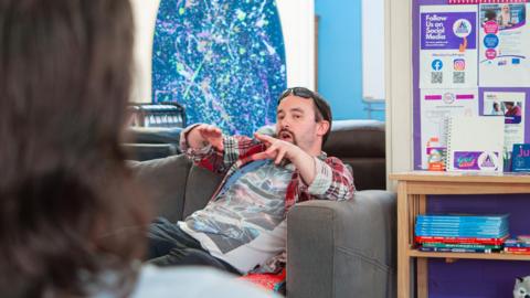 Simon is sitting on a sofa. He has a pair of sunglasses on his forehead. He is wearing a red, white and black check shirt over a t-shirt with a stylised New York skyline on it.