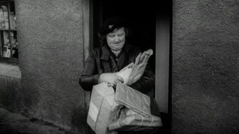 Black and white. A postwoman, Mrs Kyle, dressed black leather jacket and hat, holding a bunch of parcels, as she stands in a doorway.