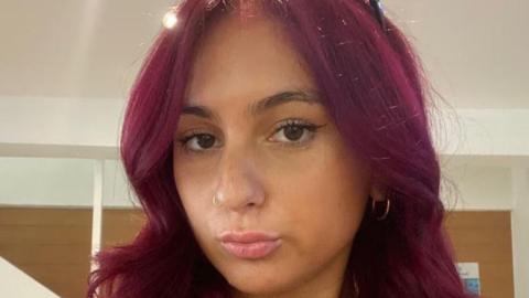 Alicia Kemp, looking at camera, with a pout. She has long hair dyed a reddish purple colour. 
