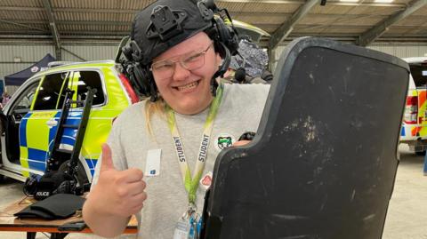 Louis is wearing a heavy-duty police helmet which has built in headphones and a camera mount at the front. He is holding a riot shield up and is smiling and giving a thumbs up to the camera. He is wearing a lanyard that says he is a student, he has blond hair and is wearing a pair of glasses. Behind him are two police cars.