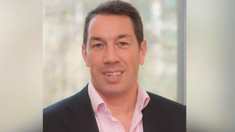 Tom Voyce in a formal photograph smiling at the camera and wearing a blue suit and pink, open-necked shirt.