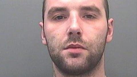 A custody image of David Braddon. He has dark stubble and looks at the camera with a neutral expression. 