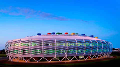 Colourful, oval-shaped, futuristic-looking Monkseaton High School which has multi-coloured blocks on its roof. The building is wrapped by white steelwork which sits on A-shaped white steel rods. Lights visible through windows make the rooms appear green coloured.