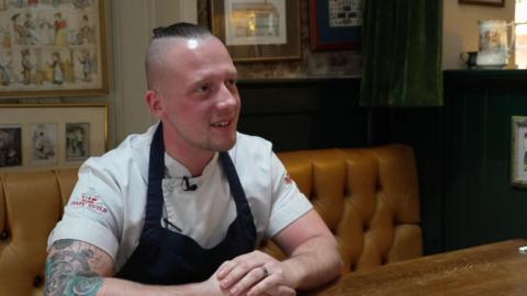 Chef Luke Emmess sitting on a table inside a pub during an interview. He is smiling and looking away from the camera.