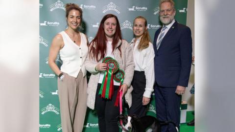 Three women and one man stand behind a black and white dog. The woman in the centre has brown hair and is holding a number of rosettes. She is also holding the dog's leash.