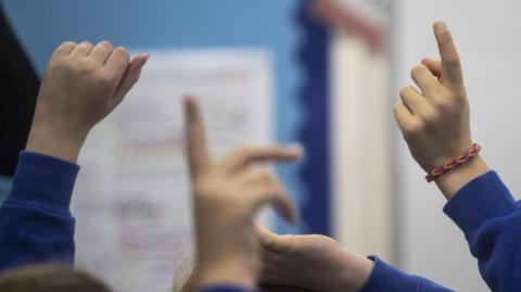 A picture of children holding their hands up in a classroom setting. They are wearing blue jumpers and one child is wearing a bracelet.