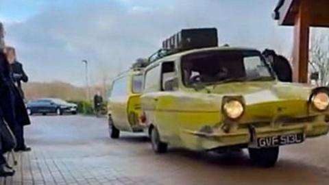 A yellow three-wheeler Robin Reliant towing a customised yellow hearse trailer.