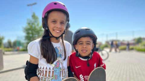 Melissa and Theo smile at the camera as they wear helmets and elbow pads. Melissa has long brown hair which comes past her shoulders and is wearing a white t-shirt with a cartoon girl by a fence on it and Theo is wearing a red Liverpool FC top. They're both holding skateboards.