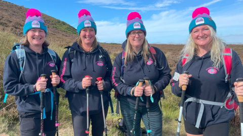 Gill Castle and three other women standing on a hill smiling into the camera. They all have light hair and are wearing blue and pink striped bobble hats, matching dark hoodies and are carrying walking poles.