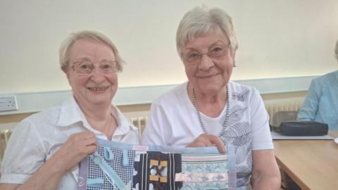 Two women with grey hair and glasses, both wearing white blouses and holding a piece of embroidery in their hands