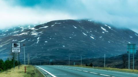 A stretch of the A9 in the Highlands. The road is single carriageway and has roadside signs warning of average speed cameras. In the background is a mountain with snow patches.
