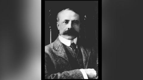 A black and white photo of Sir Edward Elgar wearing a grey suit. He has swept back short hair and a long, thick moustache.