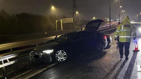 A photo of a black Audi car with its boot open and next to the central reservation of the M6. A police officer is stood next to it