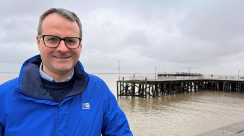 Councillor Mark Ieronimo, wearing a blue jacket and spectacles, standing next to a pier surrounded by water. The sky is grey and overcast.