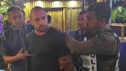 Uniformed Royal Thai Police officers arrest Ben Ransome in a restaurant, which has bamboo on the walls. He is wearing a black T-shirt and is biting his lip.