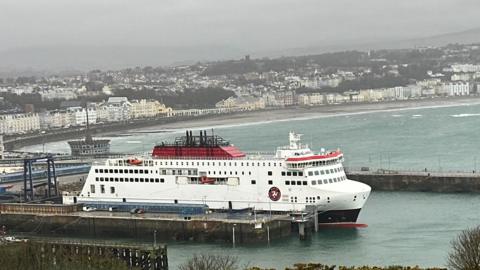 The Manxman ferry, which is white, red and black, moored in Douglas Harbour on an overcast day with Douglas Promenade in the background.