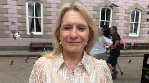 Senator Elaine Millar is wearing a cream and light brown blouse. She is standing in front of a pink and brown building and a camera crew can be seen behind her, with a TV camera on a tripod.