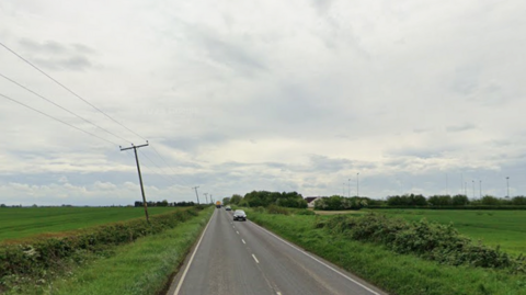 A quiet country going through open green fields, bordered by hedgerows and distant trees. Power lines trace the roadside, and a few cars travel along the tarmac under a cloudy, grey sky.