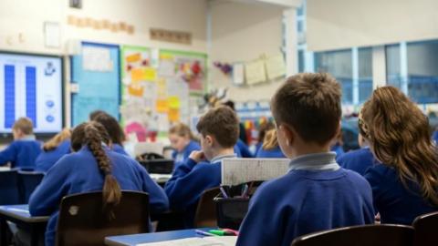A group of year five pupils sat down facing the front of a classroom. The students are wearing blue jumpers and blue polo shirts and none of their faces are visible.