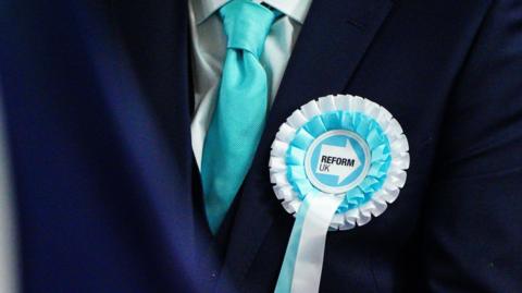 A Reform UK candidate standing with a blue and white rosette and blue tie.