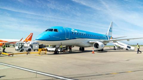 A blue KLM branded plane is seen on the ground at an airport runway, with a white and orange EasyJet plane in the background and airport workers in high vis jackets walking by.