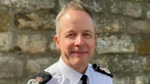 A man wearing a white shirt with a black tie and police lapels. He has short grey hair and is standing in front of a grey stone wall.