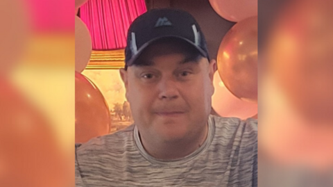 The late William Hunter looking at the camera.  He is wearing a black baseball cap and a grey t-shirt.  There are coloured balloons in the background
