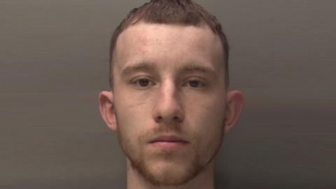 Mugshot of Jordan Cattell, who is staring into the camera with a straight face. He has short brown hair and is standing against a plain brown background