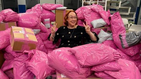 Rachel Grady sits among a large pile of pink parcels. She has a ginger curled bob and holds two peace signs up to either side of her head. She smiles widely and wears pink glasses. 