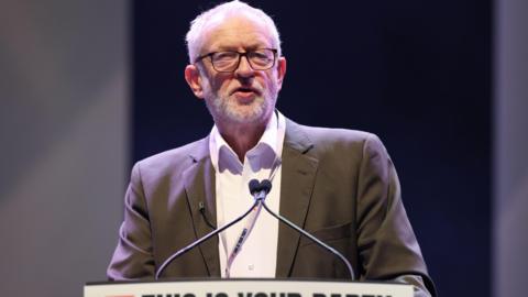 Jeremy Corbyn wearing a suit and shirt stands at a lectern addressing delegates during the Your Party conference in Liverpool