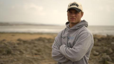 Harri Morgan standing with his arms crossed, wearing a cap and grey hoodie on the beach.