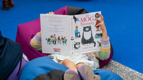A child lies on a fuchsia bean bag. She has a stripey jumper, white and blue striped dress, and shoes on as she reads The Mog Treasury - the book is completely obscuring her head. She is resting her feet on a blue bean bag. The bean bags are on a floral rug, which has been placed on a bright blue carpet.