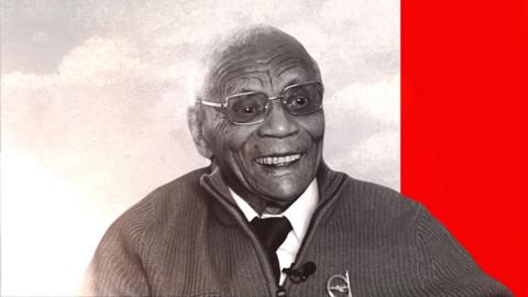 An elderly man wearing glasses and a cardigan looks past the camera smiling