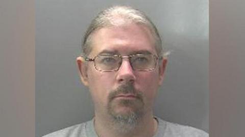 Robert Norman is looking at the camera in a police custody shot. He has fair hair that appears to be tied back, and a beard and moustache. He is wearing spectacles and a grey top.