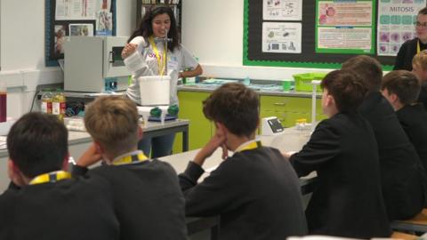 A workshop underway with a woman conducting an experiment by pouring a liquid into a bucket as students watch on