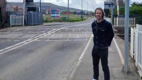 Tristan Learoyd is a man in his 30s wearing jeans. He is standing by a level crossing with a housing development behind him in the distance 